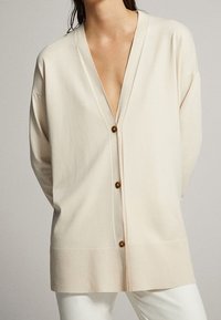 Frau trägt eine lange, hellbeige Strickjacke mit drei braunen Knöpfen über weißer Hose, stehend vor einem schlichten neutralen Hintergrund.