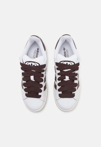 Basketball sneakers blancs avec des rayures brunes et des lacets bruns. Le matériau semble synthétique avec une texture lisse. Le logo est visible sur la languette.