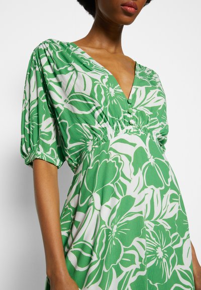 Vestido floral verde com decote em V profundo, mangas curtas com volume, tecido texturizado e padrões de flores brancas com detalhe de botões na cintura.