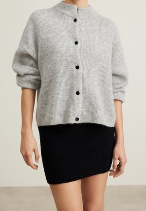Kvinde iført lysegrå cardigan med knapper og lange ærmer samt sort tekstureret mininederdel, stående mod en ensfarvet baggrund.
