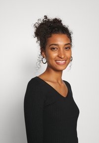 Haut à manches longues noir à côtes avec un décolleté en V. Le modèle a des cheveux bouclés et porte de grandes boucles d'oreilles. Fond blanc.
