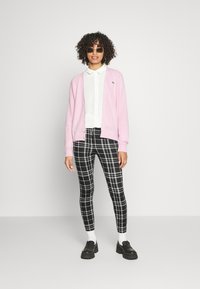 Cardigan rosa, camisa branca, calças xadrez em preto e branco, sapatos robustos pretos e óculos de sol. Cabelo encaracolado, braços relaxados ao lado.
