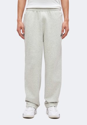 Homme portant un pantalon de survêtement gris clair et des baskets blanches, debout avec les mains relâchées le long du corps sur un fond uni.