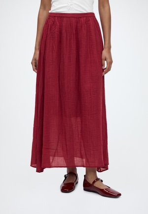 Femme portant une jupe rouge longueur cheville en tissu texturé et des chaussures rouges Mary Jane assorties, debout devant un fond uni.