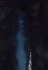 Tissu en dentelle noire superposé recouvrant partiellement un matériau en jean bleu avec une couture visible et un bord de poche en dessous.