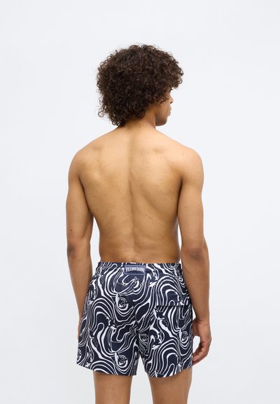 Tumši zila un balta peldbikšu komplekts ar viļņveida rakstu, izgatavots no viegla materiāla. Satur elastīgu jostasvietu un aizmugurējo kabatu.