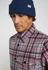 Chemise en flanelle à carreaux rouges et navy avec des boutons noirs et une poche poitrine ; tissu texturé. Bonnet en tricot bleu avec une étiquette indiquant "PULLIN".