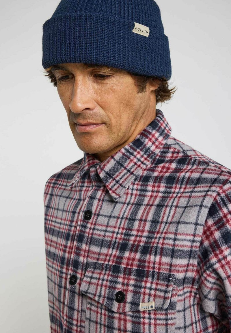 Chemise en flanelle à carreaux rouges et navy avec des boutons noirs et une poche poitrine ; tissu texturé. Bonnet en tricot bleu avec une étiquette indiquant "PULLIN".