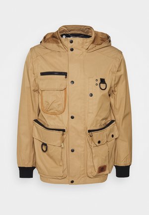 Khakifarbene Kapuzenjacke mit mehreren Reißverschluss- und Knopftaschen, schwarzen Druckknöpfen, elastischen Bündchen und schwarzen Ringen an den Vordertaschen.