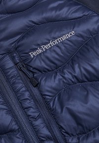 Navy blue insulated jacket with padded design, featuring a zip closure and the "PeakPerformance" logo embroidered on the chest.