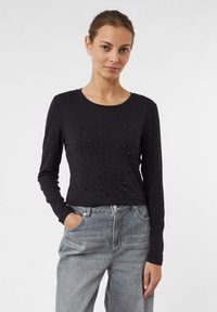 Schwarzes Longsleeve-Oberteil mit Rundhalsausschnitt, das auf der Vorderseite mit strukturierten Verzierungen versehen ist. Kombiniert mit grauen, hoch taillierten Jeans.
