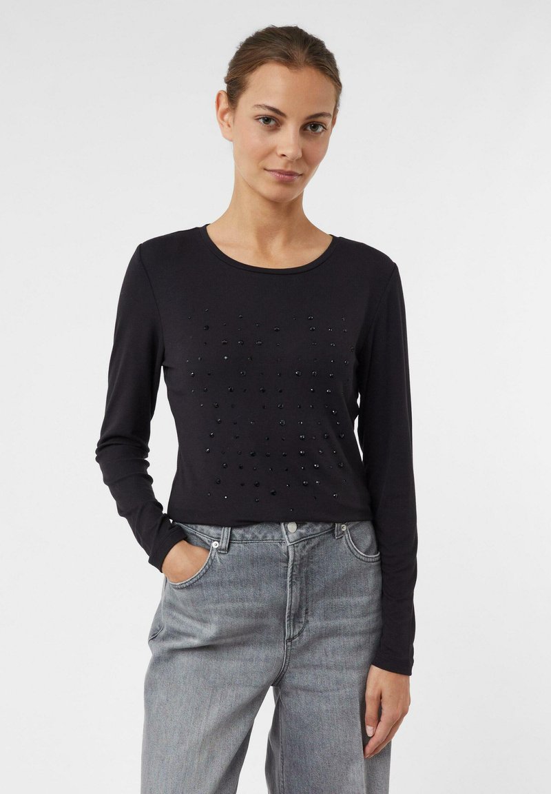 Schwarzes Longsleeve-Oberteil mit Rundhalsausschnitt, das auf der Vorderseite mit strukturierten Verzierungen versehen ist. Kombiniert mit grauen, hoch taillierten Jeans.