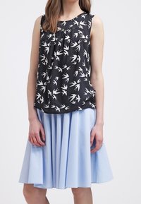 Top senza maniche nero con motivo di uccelli bianchi, abbinato a una gonna plissettata blu chiaro. Il tessuto ha una consistenza liscia e un design fluido.