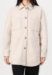 Beige long-sleeve buttoned wool jacket with two large front chest pockets and side pockets, worn over black pants.