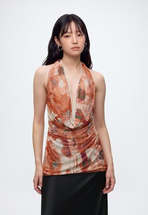 Jeune femme aux longs cheveux foncés porte un haut sans manches orange à motifs avec des strass et une jupe noire, debout devant un fond blanc.
