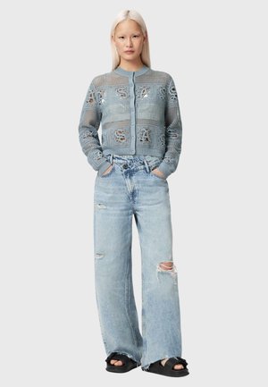 Woman with long blonde hair wearing a blue mesh buttoned cardigan, light ripped wide-leg jeans, and black slide sandals, standing with hands in pockets.