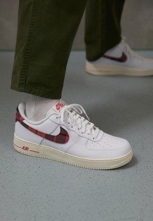 Weiße Nike Air Sneaker mit rotem kariertem Swoosh-Logo, getragen mit weißen Socken und grünen Hosen auf einem grauen, gesprenkelten Boden.