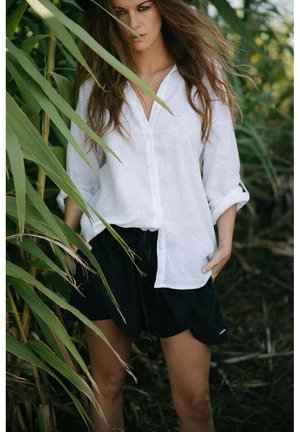 Kobieta z długimi włosami w białej koszuli zapinanej na guziki i czarnych szortach stoi częściowo za wysokimi zielonymi roślinami na zewnątrz.