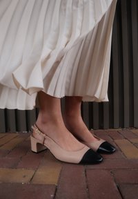Décolleté beige con punta nera, tacco quadrato e accento con fibbia dorata. Tessuto bianco a pieghe si adagia delicatamente intorno alle scarpe.