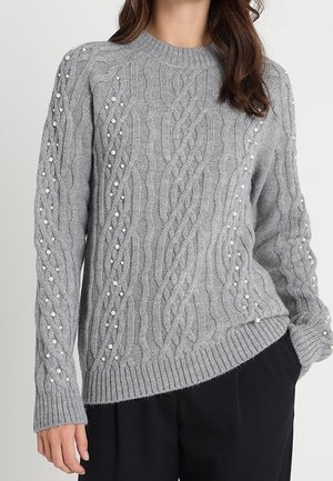 Grå kabelstrikket sweater med perleudsmykninger båret af en person med den ene hånd i lommen på sorte bukser.
