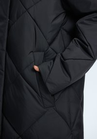 Schwarze Pufferjacke mit gestepptem Muster, ausgestattet mit einer Seitentasche. Hergestellt aus synthetischem Material, das eine glatte, matte Textur aufweist.