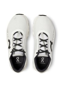 Zapatillas deportivas blancas con una parte superior texturizada, cordones negros y una suela negra y gris. Presentan un logotipo destacado y el nombre del modelo a los lados.