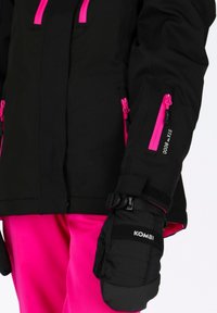 Giacca da sci nera con zip rosa, dotata di molteplici tasche e polsini regolabili. Abbinata a pantaloni rosa e muffole nere con l'etichetta "KOMBI."