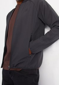 Černá sportovní bunda vyrobená z měkké, hladké látky, s vý škovým límcem, zipovými kapsami a žebrovanými akcenty podél rukávů.