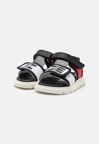 Sandalias en blanco y negro con correas de velcro, acentos rojos y una suela de goma texturizada, que presentan un logotipo distintivo en la correa.