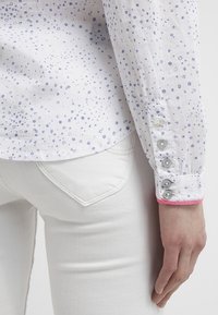 Weißes Hemd mit kleinen blauen Punktmustern, mit einem pinken Akzent an den Manschetten und silbernen Knöpfen an den Ärmeln. Kombiniert mit weißen Jeans.