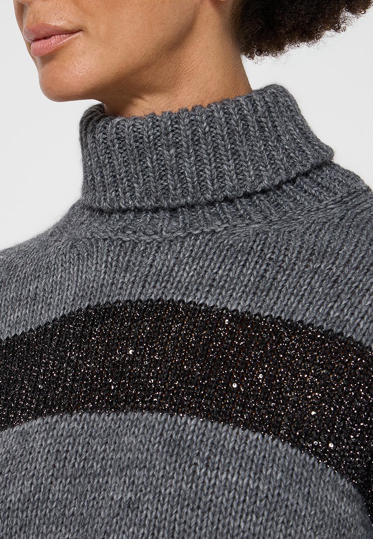 Donna che indossa un maglione grigio a collo alto lavorato a maglia con una larga striscia nera di paillettes sul petto.