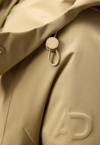 Beżowa tkanina z wytłoczonym logiem "AD" oraz okrągłym zapięciem przypiętym do pętli sznurkowej na elemencie odzieży.