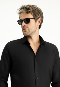 Camicia nera con bottoni realizzata in un tessuto testurizzato, con colletto a punta e pattina frontale. Indossata con occhiali da sole scuri, che mostrano uno stile contemporaneo.