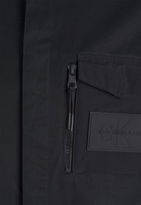 Giacca nera con tasca con zip, tessuto strutturato e patch con logo. Il design include cuciture verticali e una zip resistente.