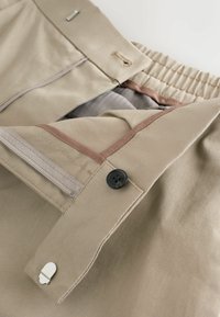 Helle Khaki-Hose mit Reißverschluss, Knopfverschluss und elastischem Bund. Sie verfügt über einen beigen Stoff mit feiner Struktur und kontrastierenden Akzenten.