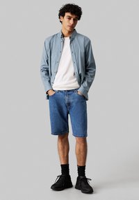 Camicia button-up a righe azzurre su una maglietta bianca, abbinata a pantaloni corti di denim. Il modello indossa sneakers nere e calze nere di lunghezza media.