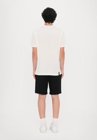 Hvit bomull t-skjorte, kort erm, minimalistisk design, sammen med svarte shorts. Modellen har på seg hvite sneakers og ankelsokker.