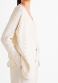 Cardigan en tricot crème avec une texture côtelée, coupe oversize et décolleté en V. Présente des épaules tombantes et une silhouette décontractée.