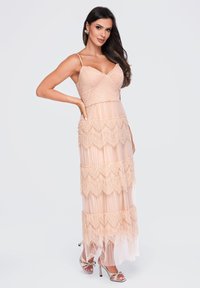 Femme aux longs cheveux foncés portant une robe maxi à volants en dentelle pêche sans manches et des sandales à talons hauts, posant avec une main sur la hanche.