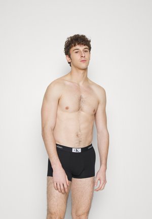 Boxer shorts de algodón negros con un logo blanco de Calvin Klein en la cinturilla. El modelo tiene el cabello rizado corto y la parte superior del cuerpo desnuda.
