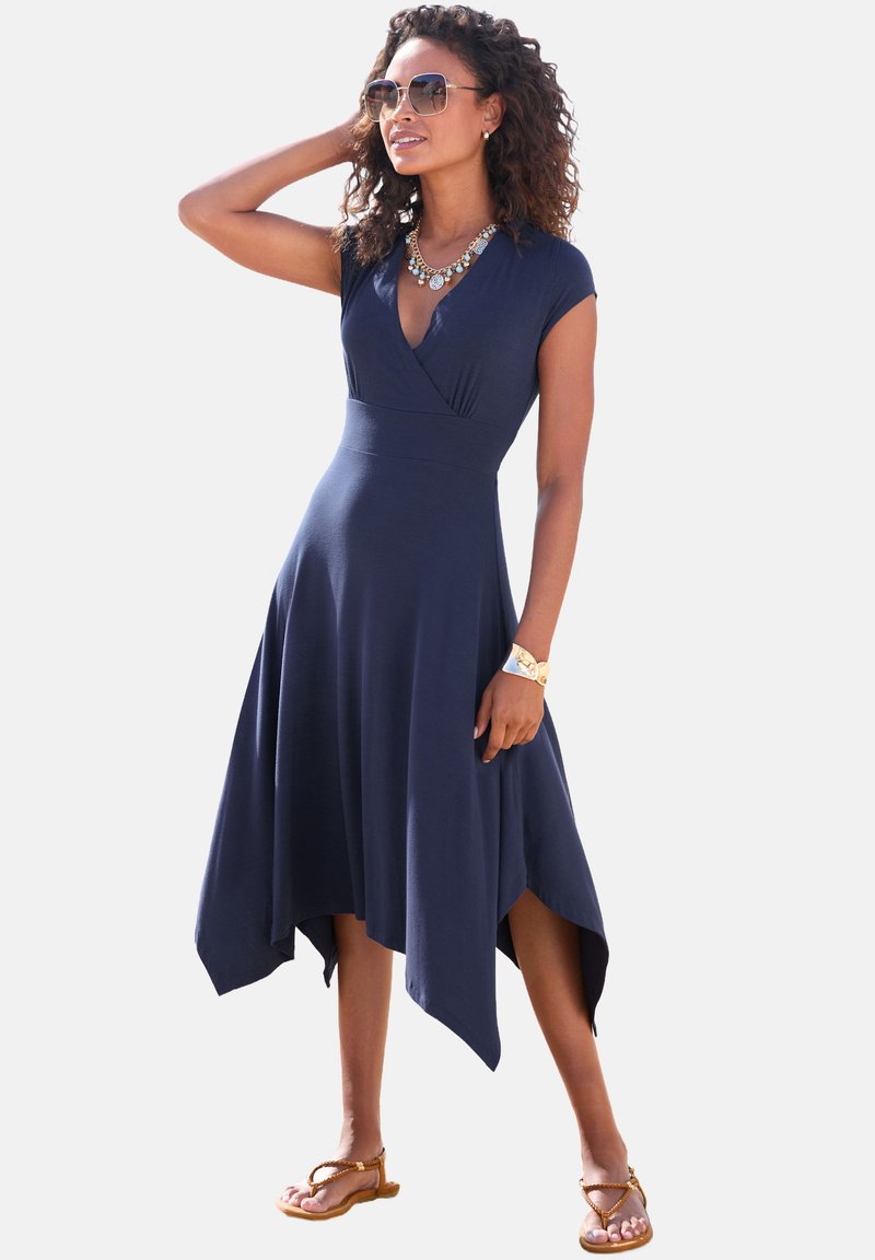 Marineblaues Kleid mit asymmetrischem Saum, kurzen Ärmeln und einem Wickel-Oberteil. Glatter Stoff, minimalistische Details, kombiniert mit Sandalen und Accessoires.