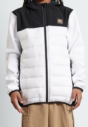 Osoba nosząca białą i czarną pikowaną kurtkę na zamek z długimi rękawami i logo ellesse na piersi, w połączeniu z beżowymi spodniami.