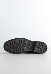 Schwarze Schuhsohle mit tiefen Profilen und kreuzförmigen Mustern, Markenname "GANT" in der Nähe des Fersenbereichs geprägt.