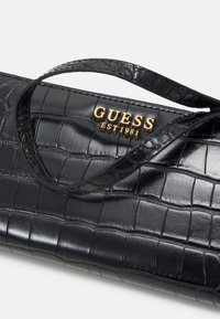 Sort syntetisk lædertaske med krokodillemønster, med et gold "GUESS EST 1981" logo og et tyndt håndtag.