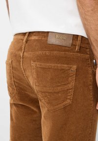 Braune Cordhosen mit einer gerippten Textur, versehen mit Gesäßtaschen und einem Leder-Brandlabel mit der Aufschrift "Brax" am Bund.
