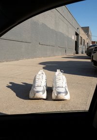 Paire de baskets blanches posée sur un trottoir en béton à côté de voitures garées et d’un mur en briques grises sous un ciel bleu clair.