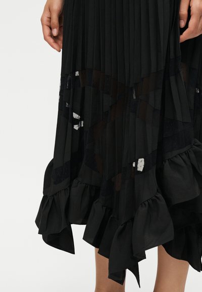 Μαύρη πλισέ φούστα με διάφανα γεωμετρικά πάνελ και βολάν στο τελείωμα, εμφανίζεται σε άτομο από μέση μηρού έως μέση, με τα χέρια να ακουμπούν στα πλευρά.