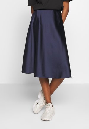 Persoon die een marineblauwe satijnen midirok, een zwart topje en witte sneakers draagt, staand met gekruiste benen tegen een effen achtergrond.