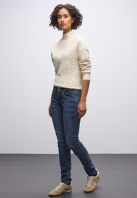 En kabelstickad, krämfärgad tröja med hög hals, tillsammans med åtsittande blå jeans och beige sneakers. Tyget verkar mjukt med en tydlig struktur.