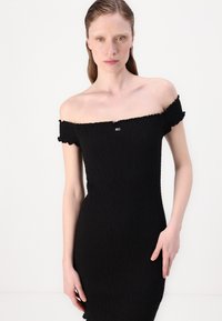 Robe noire, décolletée épaules nues, en tissu texturé, avec des manches volantes et un petit logo en accent. Silhouette ajustée.